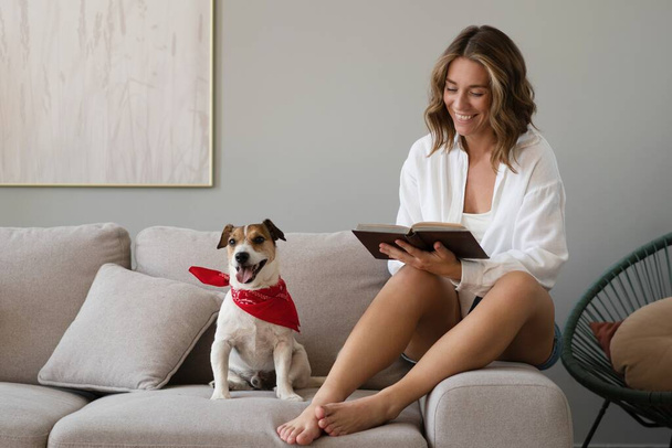 Νεαρή γυναίκα διαβάζει βιβλίο και το χαριτωμένο σκυλί της στον καναπέ στο σπίτι. Υπέροχο κατοικίδιο. - Φωτογραφία, εικόνα