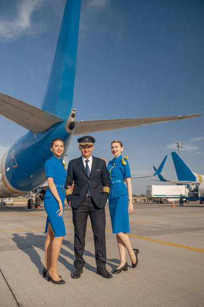 In voller Länge aufgenommen von einem fröhlichen Piloten und zwei hübschen Stewardessen, die zusammen vor einem Flugzeug stehen und in die Kamera lächeln - Foto, Bild