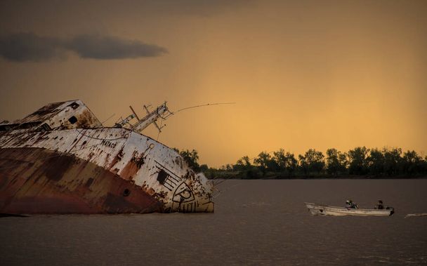 川の大きな沈没船"Mataco"小さなボートが隣に沈み、オレンジ色の空で日没時にアルゼンチンのエントラロスのParanacito, Paran River, Entre Ross, Argentina - 写真・画像