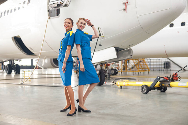 Πλήρης εικόνα δύο όμορφων αεροσυνοδών με γαλάζια στολή να χαμογελούν στην κάμερα, ποζάροντας μπροστά σε επιβατικά αεροσκάφη στο υπόστεγο του αεροδρομίου - Φωτογραφία, εικόνα