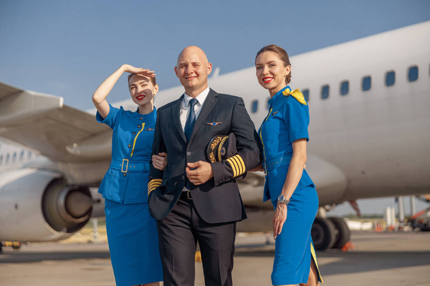 Glücklicher Pilot und zwei attraktive Stewardessen, die zusammen vor einem Flugzeug stehen und nach der Landung lächeln - Foto, Bild