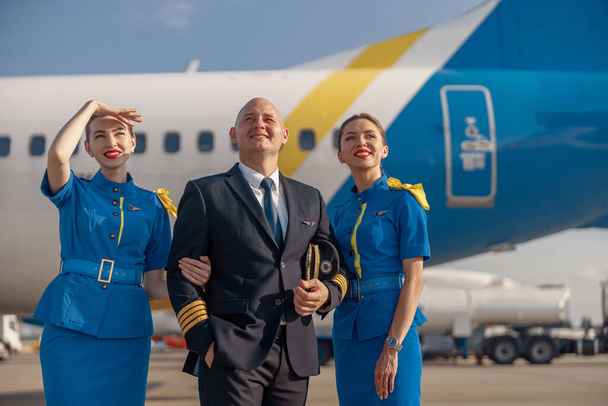 Feliz piloto y dos bellas azafatas de pie juntos frente a un avión y sonriendo después de aterrizar - Foto, imagen