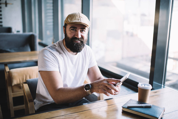 Happy bearded man in cap using smartphone while sitting at modern cafe with coffee and notebook on wooden table and looking at camera - Foto, immagini