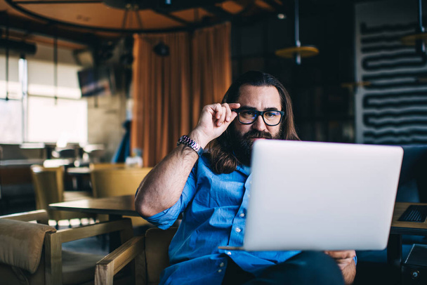Confident bearded man in glasses looking at laptop and thinking about job and sitting  on armchair at contemporary cafe with modern interior - Photo, Image