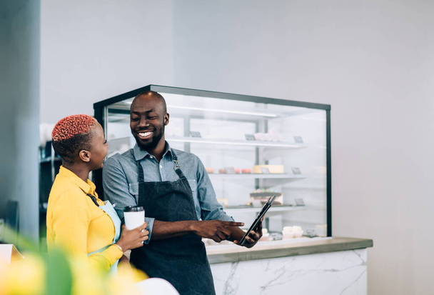 Joyful African American man pointing on digital tablet screen and laughing while standing and showing content to colleague with hot drink to go against blurred counter and showcase with desserts in light modern pastry shop - 写真・画像