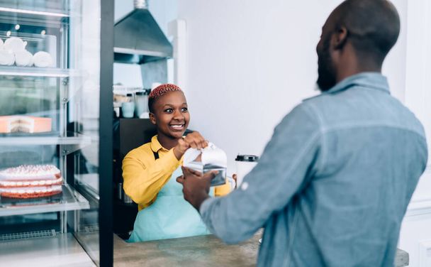 Adult African American female employee in yellow shirt and blue apron smiling and giving takeaway hot drink and food to crop blurred customer while working at counter in light modern pastry shop - Photo, image