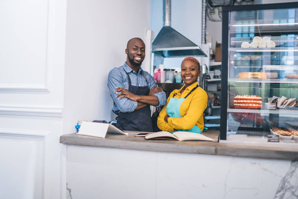 Enthusiastic active African American couple in yellow and gray shirts and blue and black aprons smiling and  looking at camera with crossed arms at counter of pastry shop - Foto, immagini