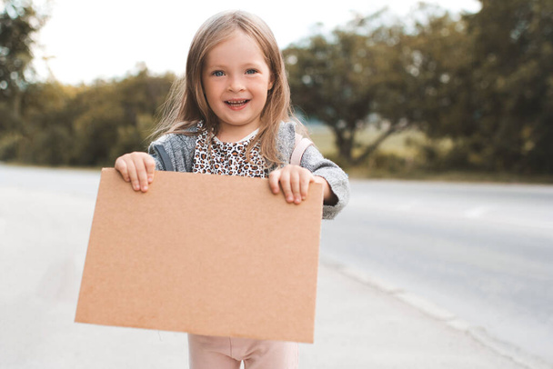 Szczęśliwy uśmiechnięty dziewczynka 3-4 lat trzymać papieru rzemiosła Paris ręcznie znak autostopem wędrówki po drogach na świeżym powietrzu. Szczęście. Małe dzieci podróżują w luźnych ciuchach i plecaku. Wakacje letnie - Zdjęcie, obraz