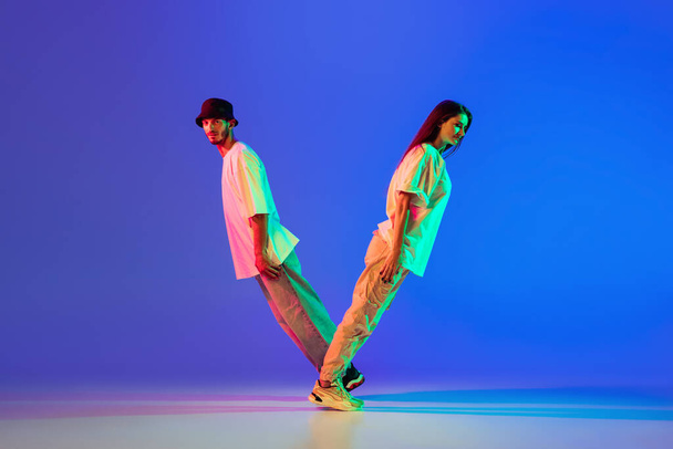 Dwoje młodych ludzi, chłopak i dziewczyna w luźnych ubraniach tańczą taniec współczesny, hip-hop na niebieskim tle w neonowym świetle. - Zdjęcie, obraz