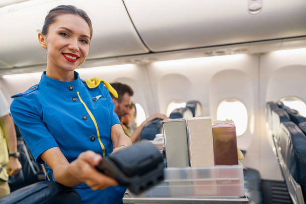 Porträt einer charmanten Stewardess, die in die Kamera lächelt und ein Terminal hält, an dem der Passagier bezahlen kann - Foto, Bild