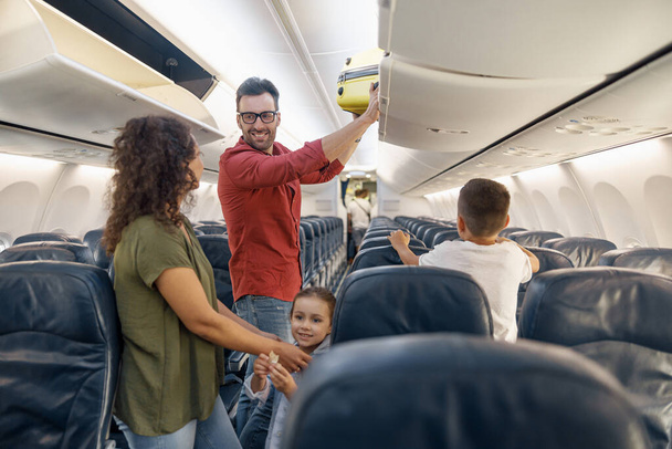 Vrolijke man die met een glimlach naar zijn vrouw kijkt en bagage in de coupé zet terwijl hij met zijn gezin met het vliegtuig reist - Foto, afbeelding