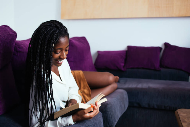 Cheerful dark skinned woman with braids smiling while read novel on free time in comfortable living room, pretty african american hipster girl lying on sofa enjoying educational literature on leisure - Photo, image