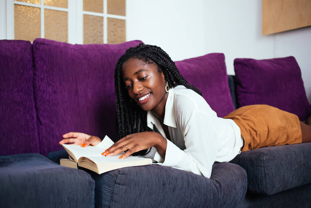 Cheerful dark skinned female in casual wear lying on sofa recreating at comfortable living room on leisure, smiling dark skinned 20s woman enjoying literature and reading interesting novel on free time - Foto, Imagem