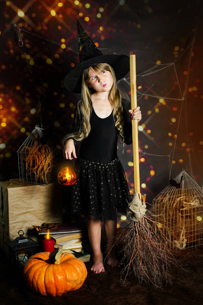 Ein Vorschulmädchen mit Hexenhut, einem Kürbis in der Hand und einem Besen mit Taschenlampe steht zwischen den Spinnweben. Halloween-Urlaubskonzept am 31. Oktober. - Foto, Bild