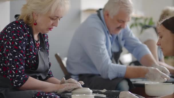Średnie ujęcie artysty ceramiki w średnim wieku uczącego grupę starszych kobiet rasy kaukaskiej i starszego mężczyzny, jak zaklinować glinę siedząc przy biurku w pracowni artystycznej. Ludzie lubią rozmawiać w pracy. - Materiał filmowy, wideo