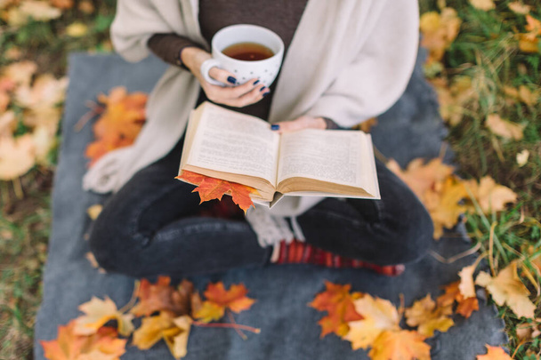 Donna in jeans neri, maglione marrone e sciarpa beige leggere libro e bere tè. Tazza con bevanda riscaldante. Foglie d'acero cadute sotto i piedi. Morbida attenzione sulla foglia all'interno del libro. Modo autunnale. - Foto, immagini