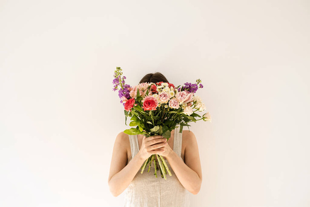 白い背景に女性の手にカラフルな花の花束。中性ベージュの若い女性はリネンのドレスを洗った顔を隠す。審美的な美しさとファッションの花のコンセプト。花の花束に隠された顔 - 写真・画像