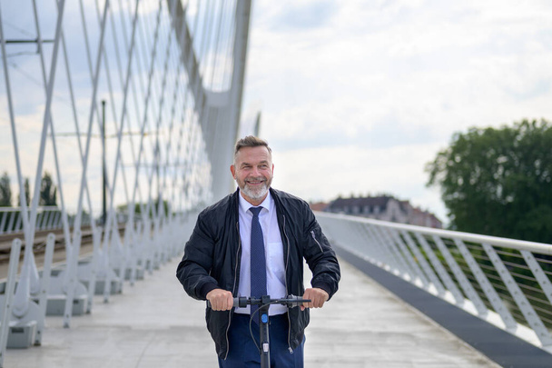 Smiling friendly businessman riding an electric scooter as he commutes home from the office at sunset crossing a long bridge over a river - Фото, изображение