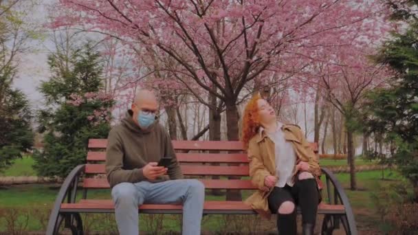 European Man with mask and woman without mask sitting on bench in park. Woman coughs contagious. Coronavirus epidemic - Footage, Video