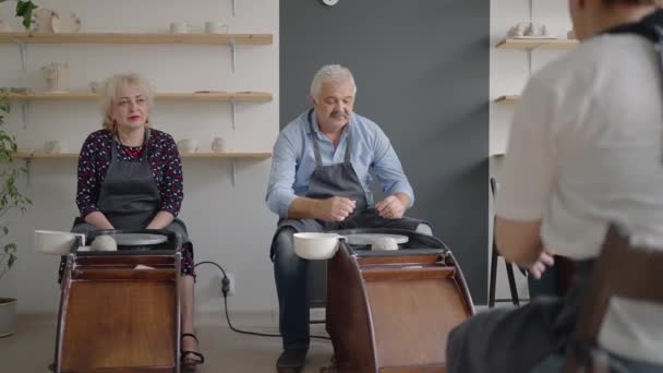 Średnie ujęcie artysty ceramiki w średnim wieku uczącego grupę starszych kobiet rasy kaukaskiej i starszego mężczyzny, jak zaklinować glinę siedząc przy biurku w pracowni artystycznej. Ludzie lubią rozmawiać w pracy. - Materiał filmowy, wideo