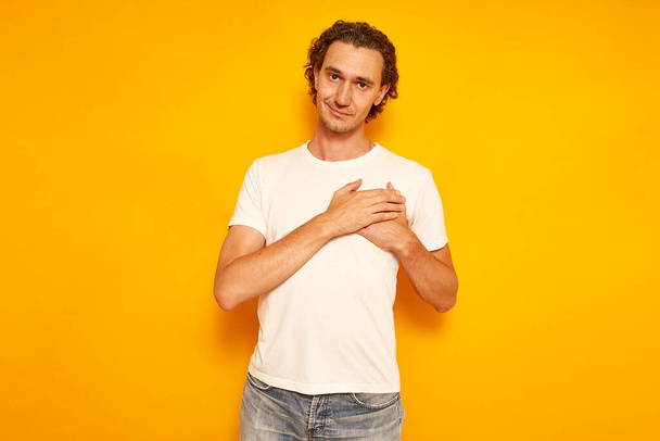 Indoor shot of handsome man keeps hands on chest, being cordial and friendly, expresses gratitude and thankfulness, has pleasant smile, dressed in casual outfit, isolated on yellow studio wall - Photo, Image