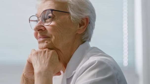 Mosolygó idősebb orvos szemüveggel támaszkodik a kezét az irodában - Felvétel, videó