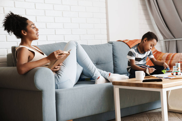 Black woman reading book while her son playing with toys at home - Foto, Bild