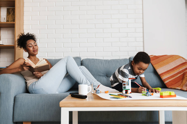 Black woman reading book while her son drawing and playing with toys at home - Photo, image