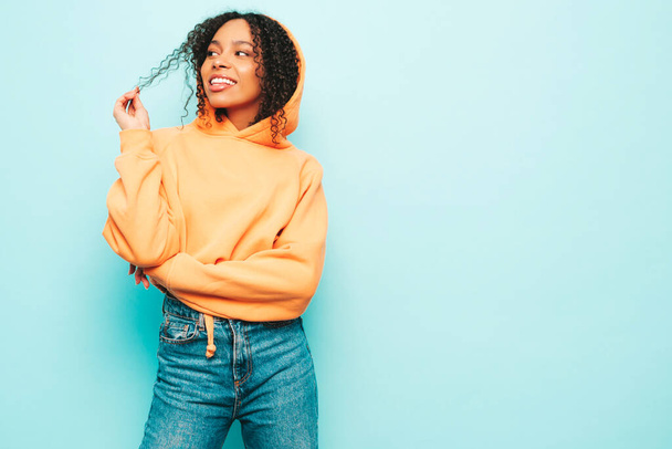 Beautiful black woman with afro curls hairstyle.Smiling model in orange hoodie and trendy jeans clothes. Sexy carefree female posing near blue wall in studio. Cheerful and happy - Фото, зображення