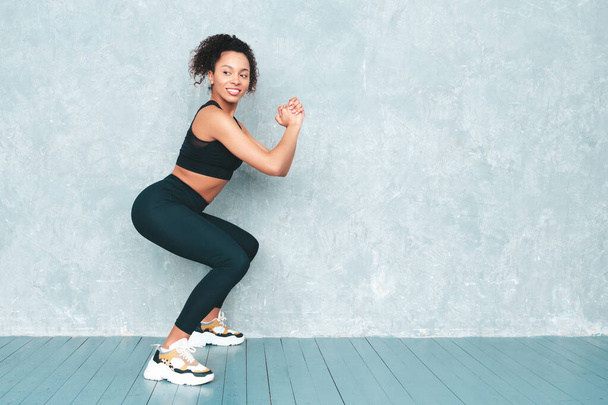 Fitness smiling black woman in sports clothing with afro curls hairstyle.She doing squats. Young beautiful model with perfect tanned body.Female in studio near gray wall. Happy and cheerful  - 写真・画像