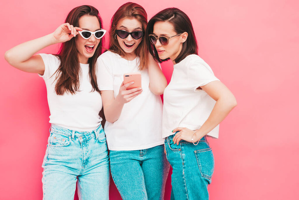 Three young beautiful smiling hipster female in trendy summer  t-shirt and jeans clothes. Sexy carefree women posing near pink wall in studio.Positive models looking at smartphone screen. Using apps  - Фото, изображение