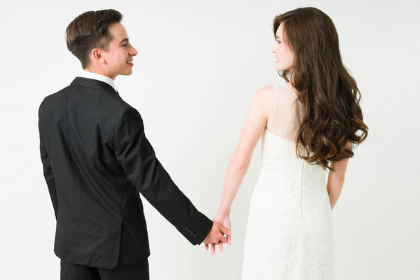 Rückansicht einer glücklichen jungen Frau im Brautkleid und eines Mannes im schwarzen Anzug, der Händchen hält  - Foto, Bild