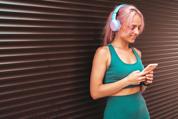 Fitness smiling woman in green sports clothing with pink hair. Young beautiful model with perfect body.Female in the street near roller shutter wall.Listening music in wireless headphones - Foto, Bild