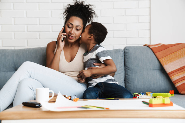 Black smiling woman talking on cellphone while hugging her son at home - Φωτογραφία, εικόνα