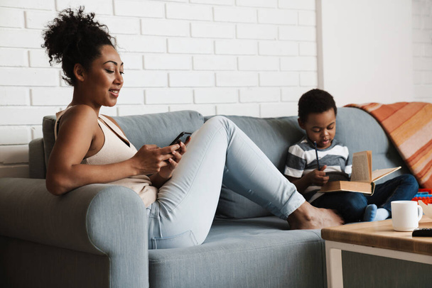 Black smiling woman using cellphone while her son reading book at home - Φωτογραφία, εικόνα