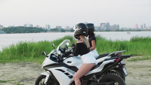 Dos mujeres amigas con cascos y sentadas en motocicletas cerca del agua cerca del centro de la ciudad y hablando - Imágenes, Vídeo