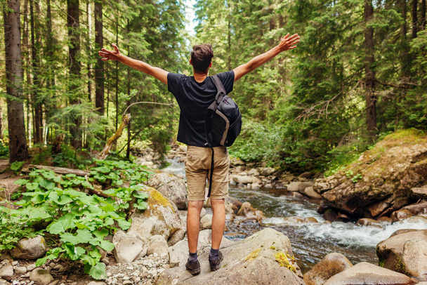 Traveler hiker with backpack raising arms by mountain river in Carpathian forest feeling happy. Man tourist enjoys landscape. Trip to summer Ukraine - Fotoğraf, Görsel