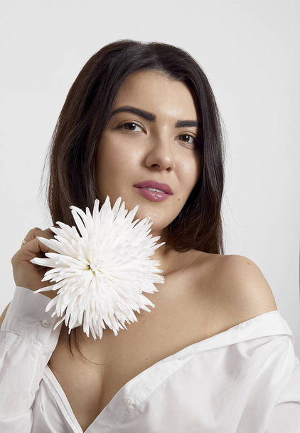 Close-up portrait of a sensual brunette young woman in a white t shirt holding a white flower, looking at camera, isolate on a white background. - Foto, Bild