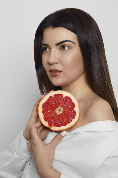 Portrait of a young woman dressed in a white t shirt holding near face grapefruit isolated on white background studio portrait. Cosmetology concept. - 写真・画像