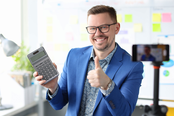Smiling blogger businessman sostiene una calculadora y graba videos de entrenamiento sobre ganancias - Foto, Imagen