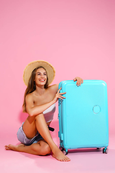 A young woman in a hat, denim shorts and white top while sitting near a blue suitcase - Photo, image