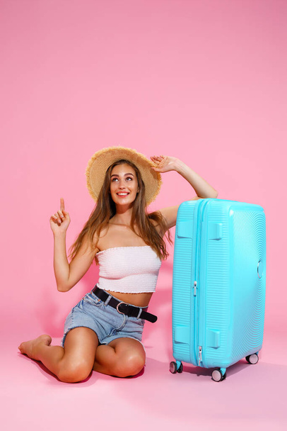 A young woman in a hat, denim shorts and white top while sitting near a blue suitcase pointing to empty space - Фото, зображення