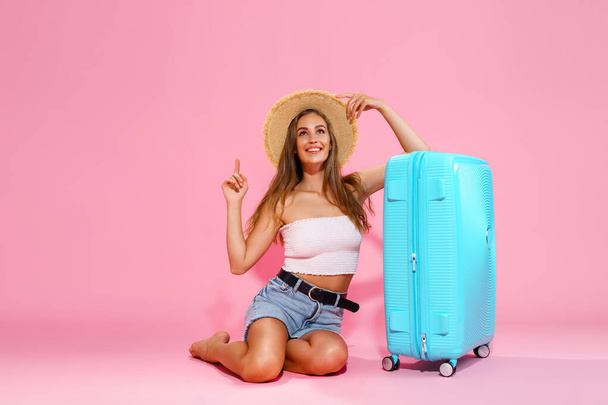 A young woman in a hat, denim shorts and white top while sitting near a blue suitcase pointing to empty space - Fotografie, Obrázek