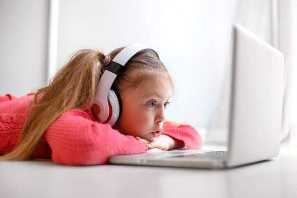 Cute little girl in headphones seriously using laptop at home for online education, home studying and online communication. Kids distance learning. Social distance. Stay at home. - Fotó, kép