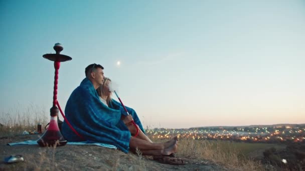 Ein Mann und ein Mädchen in den Bergen schauen den Sonnenuntergang an, in eine Decke gehüllt, sie rauchen eine Wasserpfeife, sitzen in einer Umarmung. Schauen Sie sich die nächtlichen Lichter der Stadt an. Ruhe in der Stadt. - Filmmaterial, Video