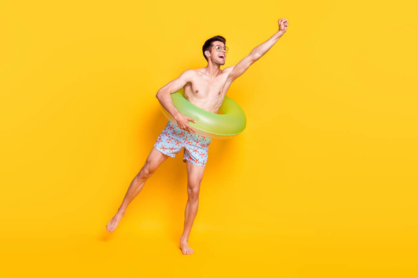 Photo of cute funky guy shirtless dressed dark glasses smiling inside buoy catching arm empty space isolated yellow color background - Zdjęcie, obraz