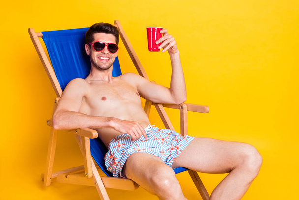 Photo of sweet charismatic young man naked torso lying lounge chair drinking beer isolated yellow color background - 写真・画像