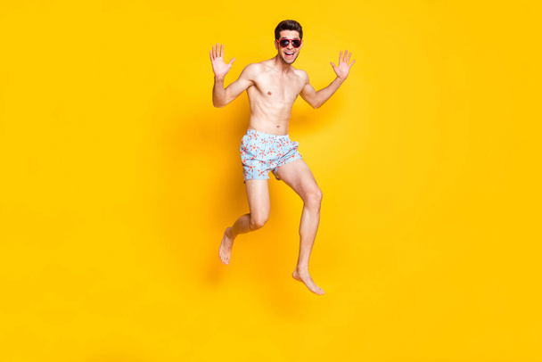 Photo of charismatic funky young man naked torso wear dark eyewear jumping high smiling isolated yellow color background - 写真・画像