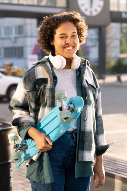 Сміється милий підліток зі скейтбордом, що стоїть у міському середовищі
 - Фото, зображення