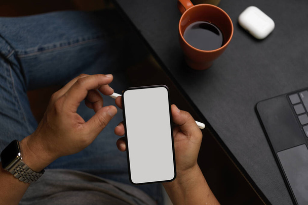 Κοντινό πλάνο, top view, Άντρας μεσήλικας χρησιμοποιώντας smartphone που εργάζονται από το σπίτι, μαύρο καφέ, tablet, ακουστικά σε μαύρο τραπέζι εργασίας, τηλέφωνο οθόνη mockup - Φωτογραφία, εικόνα
